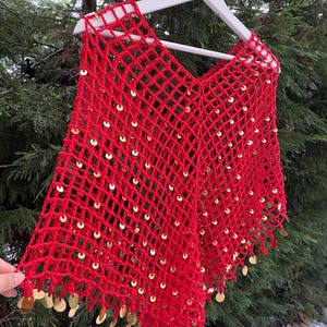 May include: A vibrant red, open-weave poncho with a V-neck design. The poncho is adorned with numerous gold sequins, adding a touch of sparkle. The edges are finished with a fringe of gold sequins. The garment is displayed on a white hanger against a backdrop of green foliage.