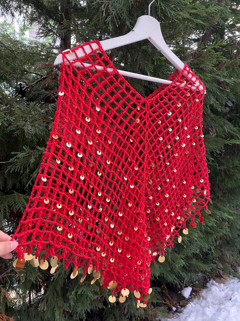 May include: A vibrant red, open-weave poncho with a V-neck design. The poncho is adorned with numerous gold sequins, adding a touch of sparkle. The edges are finished with a fringe of gold sequins. The garment is displayed on a white hanger against a backdrop of green foliage.