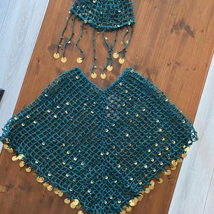 May include: A teal green crocheted outfit, including a poncho and a head covering. The poncho and head covering are adorned with gold-colored circular sequins. The head covering has dangling strands with gold sequins. The outfit is displayed on a wooden surface.