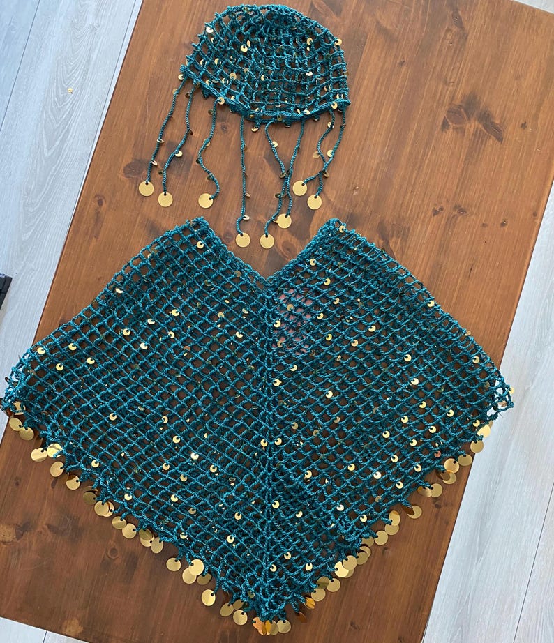 May include: A teal green crocheted outfit, including a poncho and a head covering. The poncho and head covering are adorned with gold-colored circular sequins. The head covering has dangling strands with gold sequins. The outfit is displayed on a wooden surface.