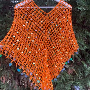 May include: An orange crochet poncho with a V-neck design, adorned with iridescent sequins. The open weave of the poncho is visible, and the sequins are attached along the edges, creating a shimmering effect. The poncho is displayed on a white hanger.