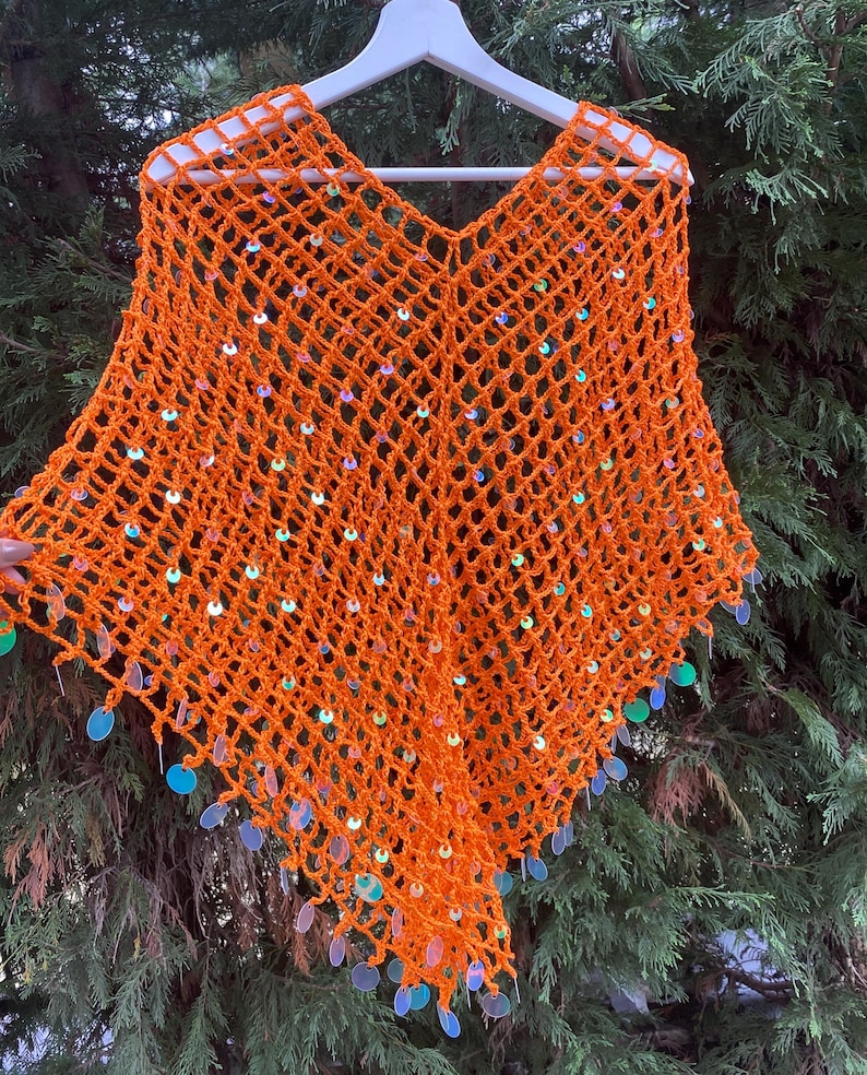 May include: An orange crochet poncho with a V-neck design, adorned with iridescent sequins. The open weave of the poncho is visible, and the sequins are attached along the edges, creating a shimmering effect. The poncho is displayed on a white hanger.