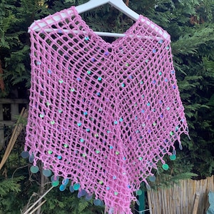 Może przedstawiać: Różowy, ażurowy poncho z szydełka z iryzującymi cekinami. Poncho jest prezentowane na białym wieszaku na tle zielonych liści. Dolna krawędź poncho jest ozdobiona wiszącymi cekinami.