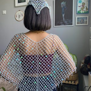 Puede incluir: Un chal de ganchillo plateado con un patrón de malla abierta en forma de diamante, adornado con lentejuelas iridiscentes. El chal tiene un dobladillo en punta y se combina con un tocado de ganchillo plateado a juego con diseño similar.