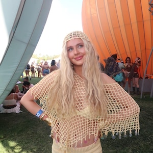 May include: A light yellow crochet outfit, including a top and shorts, worn with a matching headband. The top has a net-like design, and the shorts are high-waisted. The outfit is accessorized with gold bracelets. The background includes an orange balloon structure.