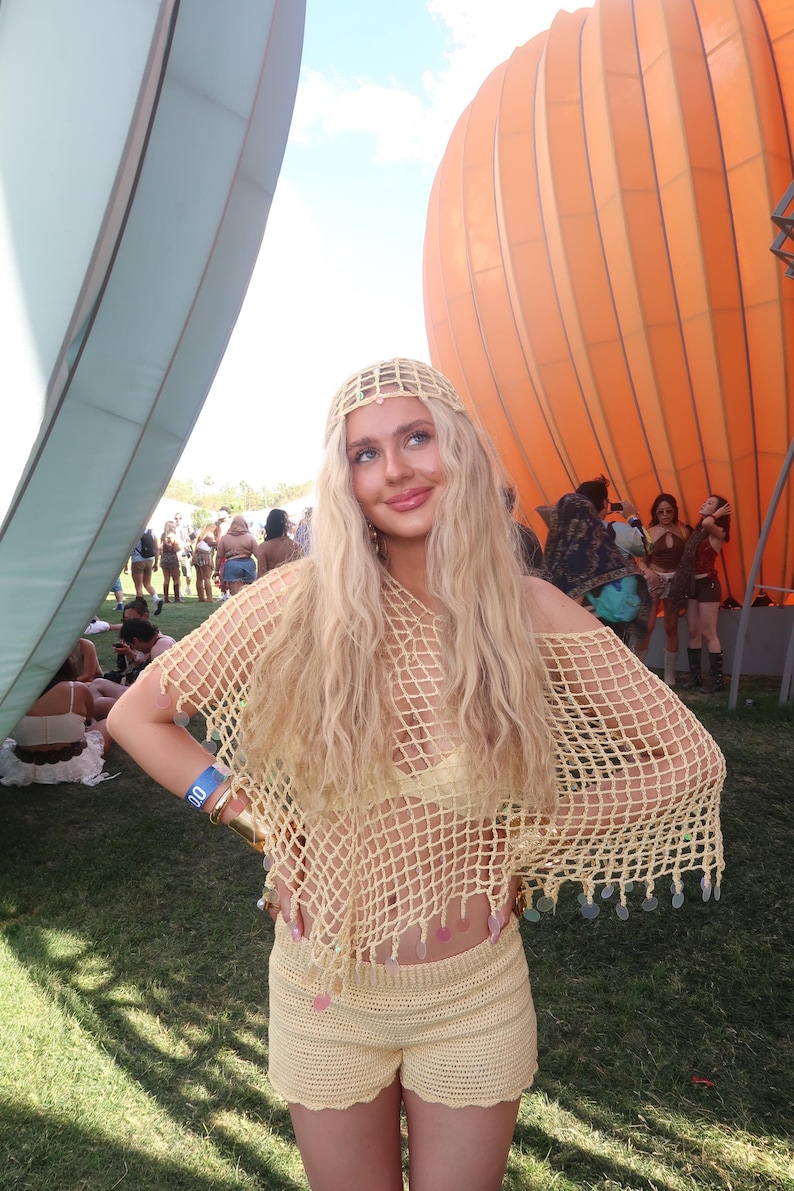 May include: A light yellow crochet outfit, including a top and shorts, worn with a matching headband. The top has a net-like design, and the shorts are high-waisted. The outfit is accessorized with gold bracelets. The background includes an orange balloon structure.