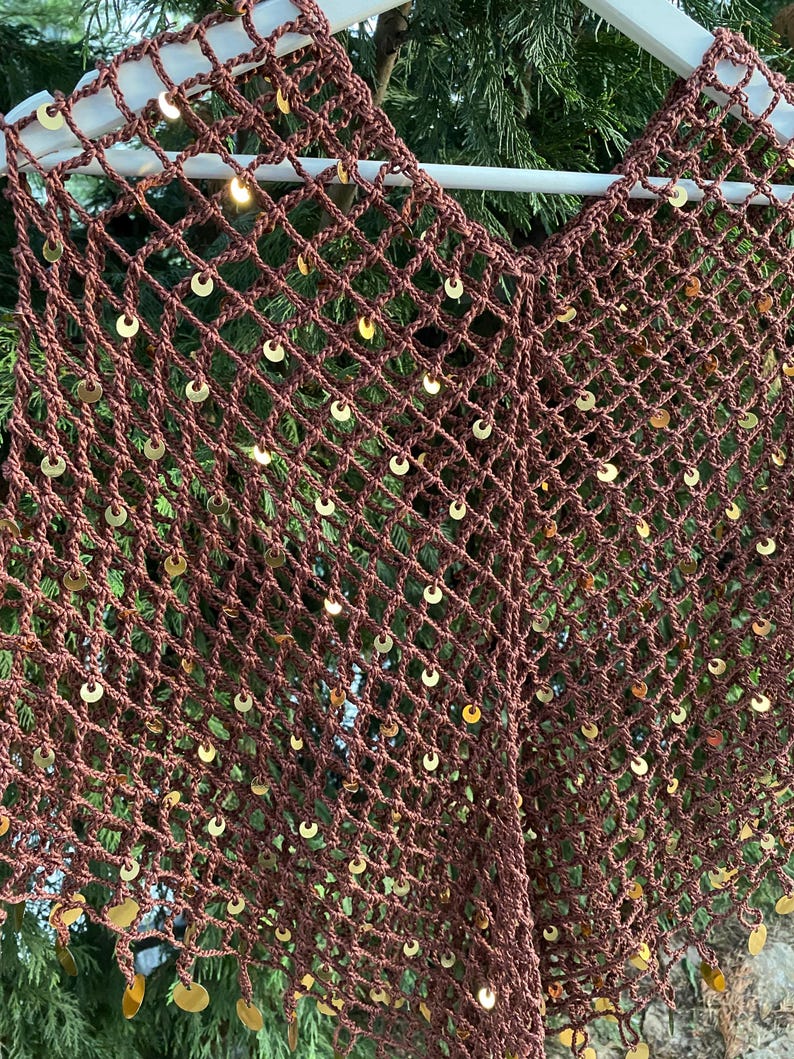 May include: A brown, open-weave crochet top with a V-neck design. The top is adorned with numerous gold-colored sequins, adding a touch of sparkle. The garment is displayed on a white hanger against a backdrop of green foliage.