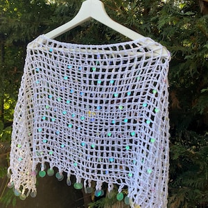 May include: White crochet shawl with iridescent sequins. The open weave design is accented with dangling, round sequins along the bottom edge. The shawl is displayed on a white hanger against a backdrop of green foliage.