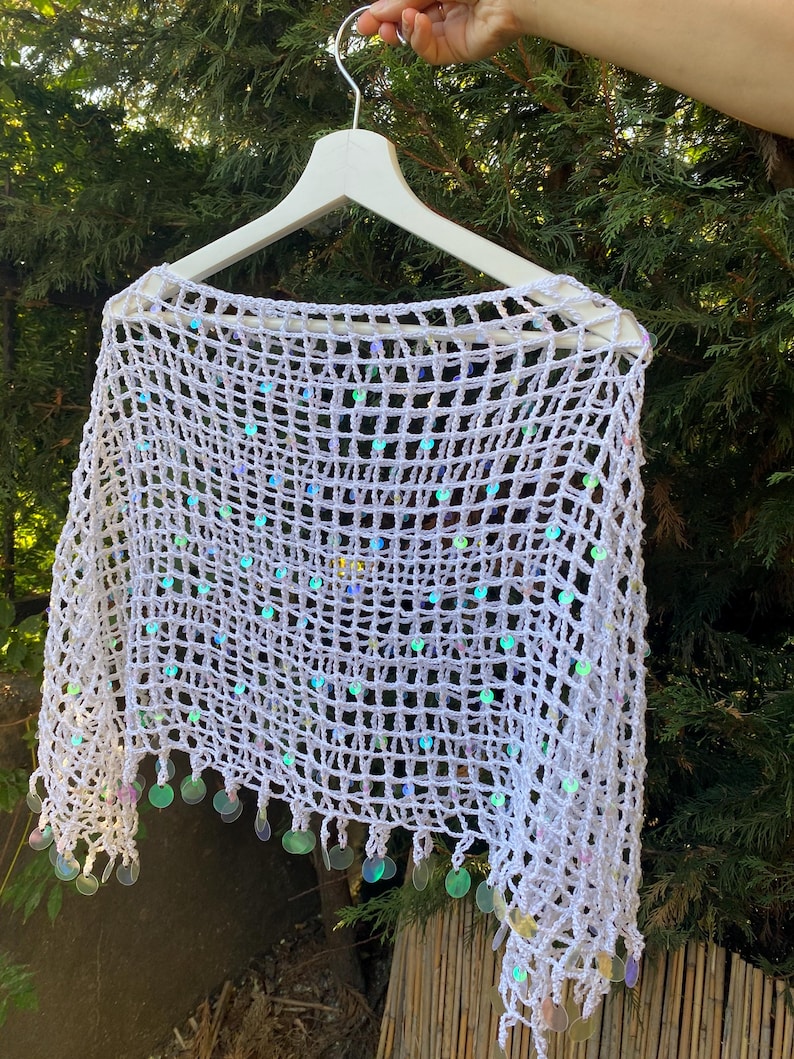 May include: White crochet shawl with iridescent sequins. The open weave design is accented with dangling, round sequins along the bottom edge. The shawl is displayed on a white hanger against a backdrop of green foliage.