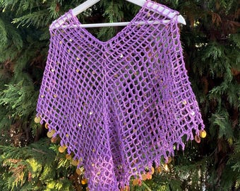 May include: A purple, open-weave poncho with a V-neck, hanging on a white hanger. The poncho is adorned with gold-colored sequins along the bottom edge. The background is a green, leafy tree.