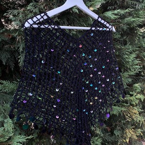 May include: Black crocheted poncho with iridescent sequins. The open weave design features a V-neck and a fringe of sequins along the bottom edge. The poncho is displayed on a white hanger against a backdrop of green foliage.