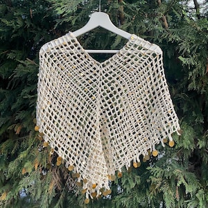 May include: Cream-colored, open-weave poncho with a V-neck design. The poncho is adorned with gold-colored sequins along the bottom edge. The garment is displayed on a white hanger against a backdrop of green foliage.