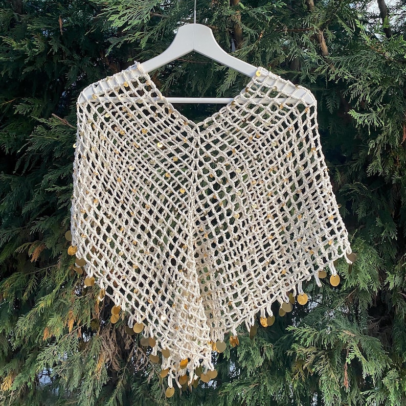 May include: Cream-colored, open-weave poncho with a V-neck design. The poncho is adorned with gold-colored sequins along the bottom edge. The garment is displayed on a white hanger against a backdrop of green foliage.