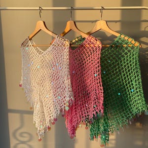May include: Three crocheted ponchos in white, pink, and green hang on wooden hangers. Each poncho has a V-neck and is adorned with iridescent sequins and dangling embellishments. The ponchos are displayed against a neutral wall.