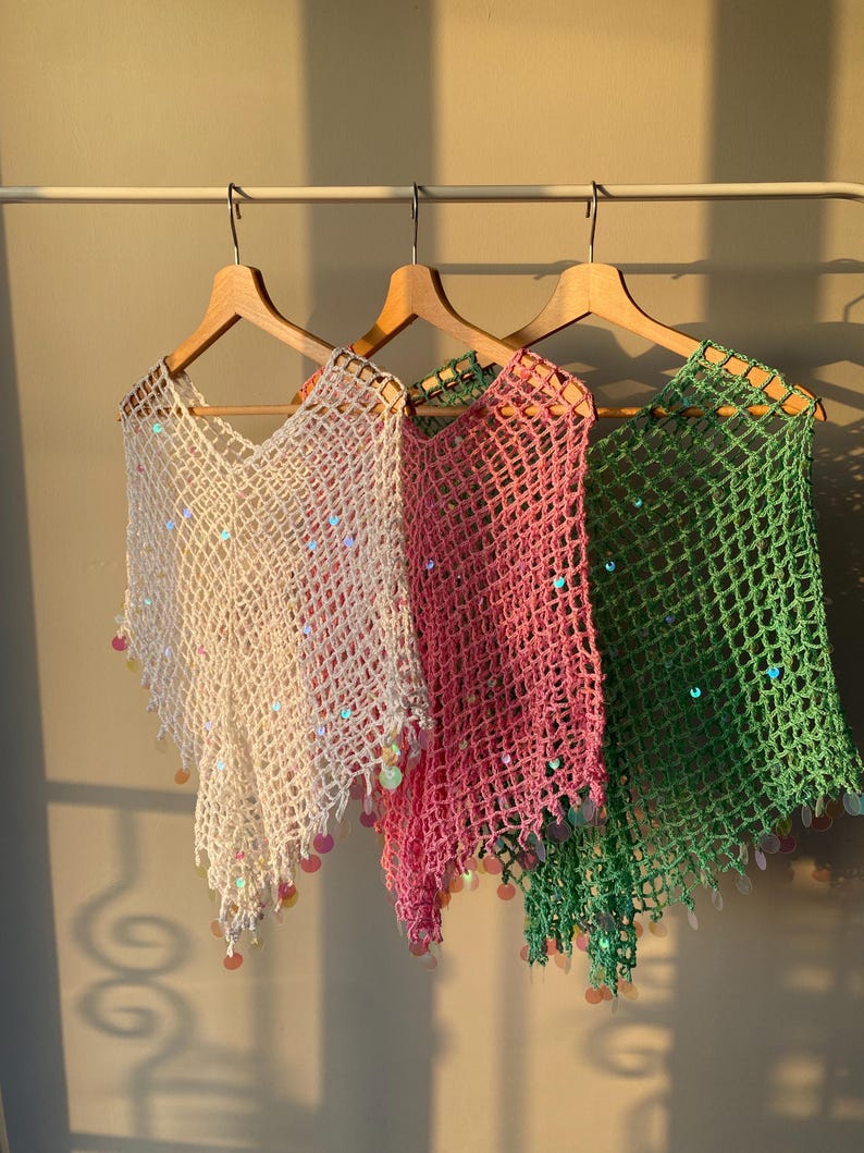 May include: Three crocheted ponchos in white, pink, and green hang on wooden hangers. Each poncho has a V-neck and is adorned with iridescent sequins and dangling embellishments. The ponchos are displayed against a neutral wall.