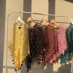 May include: A collection of open-weave ponchos in various colors, including yellow, black, brown, pink, and teal, hang on a white rack. Each poncho is adorned with gold-colored sequins along the bottom edge, creating a shimmering effect.