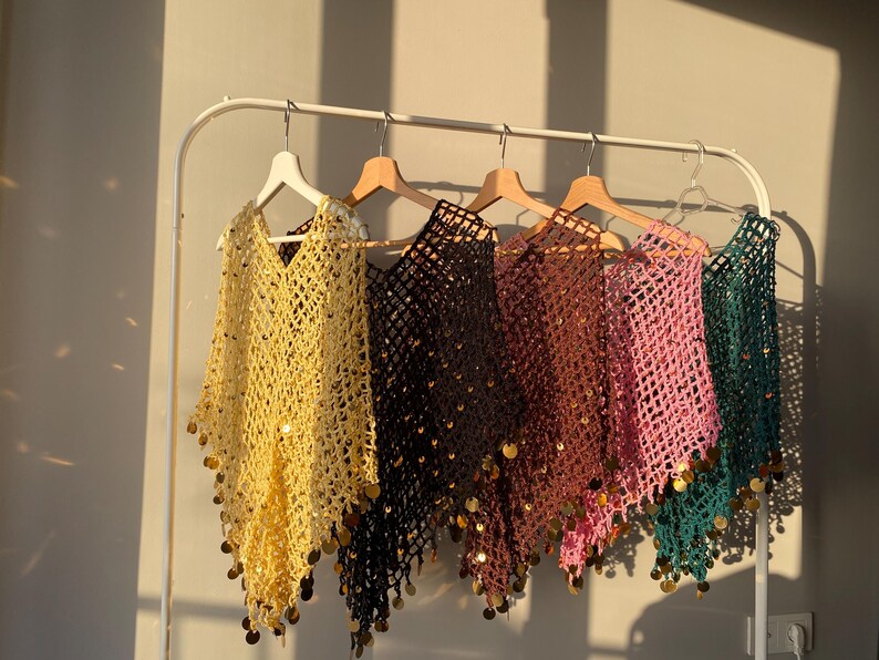 May include: A collection of open-weave ponchos in various colors, including yellow, black, brown, pink, and teal, hang on a white rack. Each poncho is adorned with gold-colored sequins along the bottom edge, creating a shimmering effect.