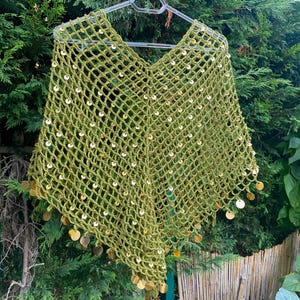 May include: A green crocheted poncho with a V-neck design, adorned with gold-colored sequins. The open-weave pattern is visible, and the poncho is hanging on a hanger against a backdrop of green foliage. The sequins are attached to the bottom edge.