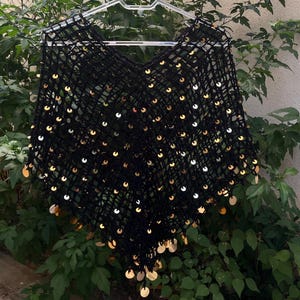 May include: A black, open-weave poncho with a V-neck, adorned with numerous gold-colored sequins. The garment is displayed on a clear plastic hanger against a backdrop of green foliage. The sequins are evenly spaced across the fabric.