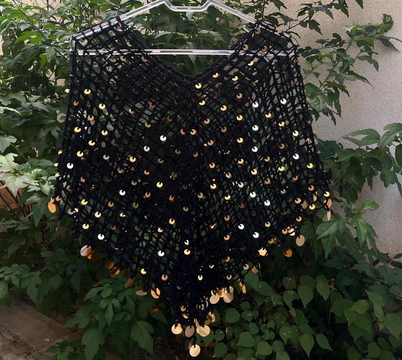May include: A black, open-weave poncho with a V-neck, adorned with numerous gold-colored sequins. The garment is displayed on a clear plastic hanger against a backdrop of green foliage. The sequins are evenly spaced across the fabric.
