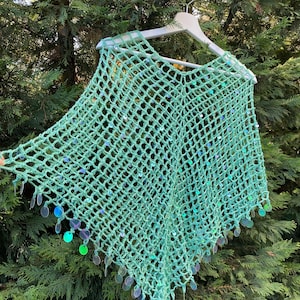 May include: A mint green crochet poncho with a net-like design, embellished with iridescent sequins. The poncho is hanging on a white hanger against a backdrop of green foliage. The sequins add a touch of sparkle to the handmade garment.