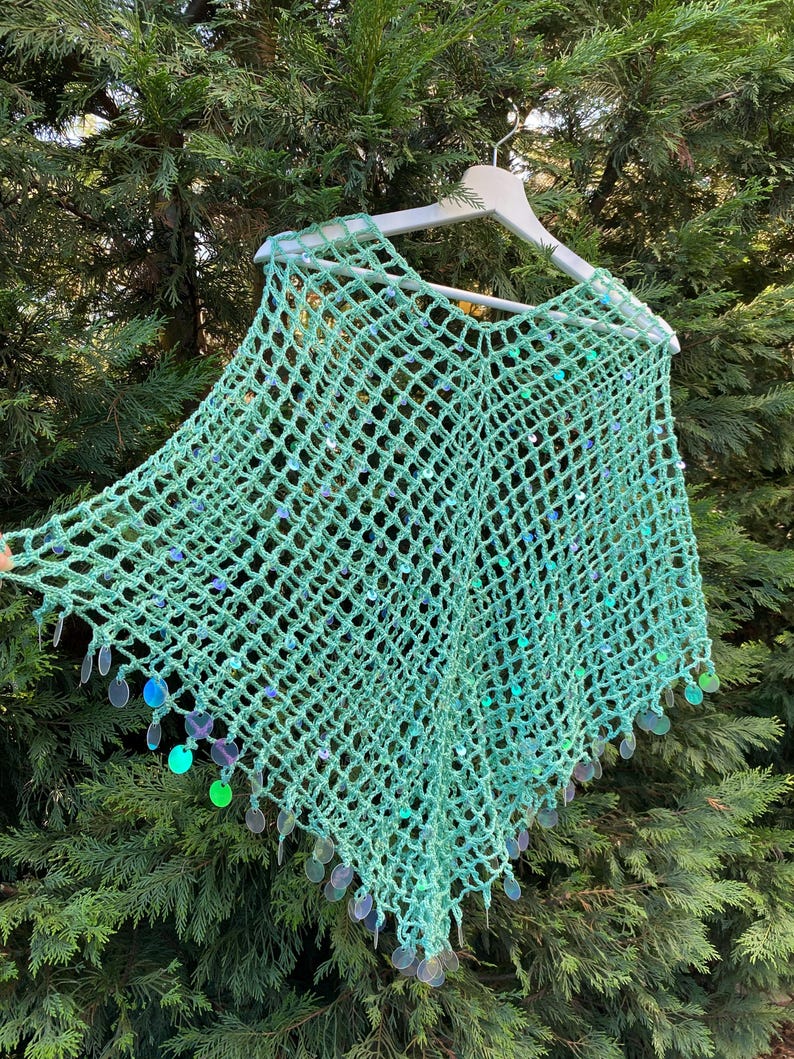 May include: A mint green crochet poncho with a net-like design, embellished with iridescent sequins. The poncho is hanging on a white hanger against a backdrop of green foliage. The sequins add a touch of sparkle to the handmade garment.