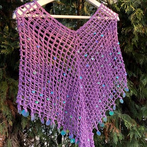 May include: A handmade, open-weave crochet poncho in a vibrant purple hue, adorned with iridescent sequins. The poncho is displayed on a wooden hanger, showcasing its delicate net-like pattern and flowing design. A unique, eye-catching garment.