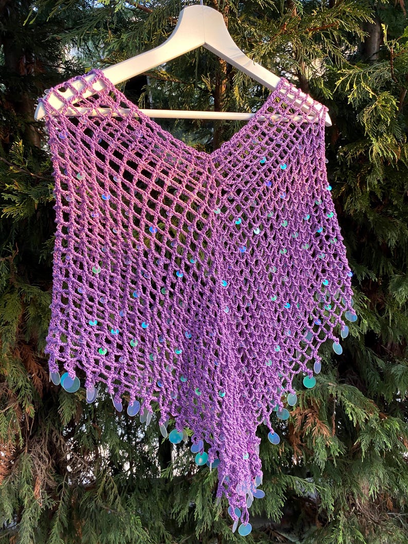 May include: A handmade, open-weave crochet poncho in a vibrant purple hue, adorned with iridescent sequins. The poncho is displayed on a wooden hanger, showcasing its delicate net-like pattern and flowing design. A unique, eye-catching garment.