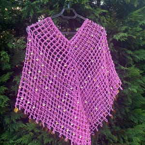 May include: A pink, open-weave crochet poncho with a V-neck. The poncho is adorned with gold-colored sequins throughout the body and along the bottom edge. The garment is displayed on a black hanger against a backdrop of green foliage.