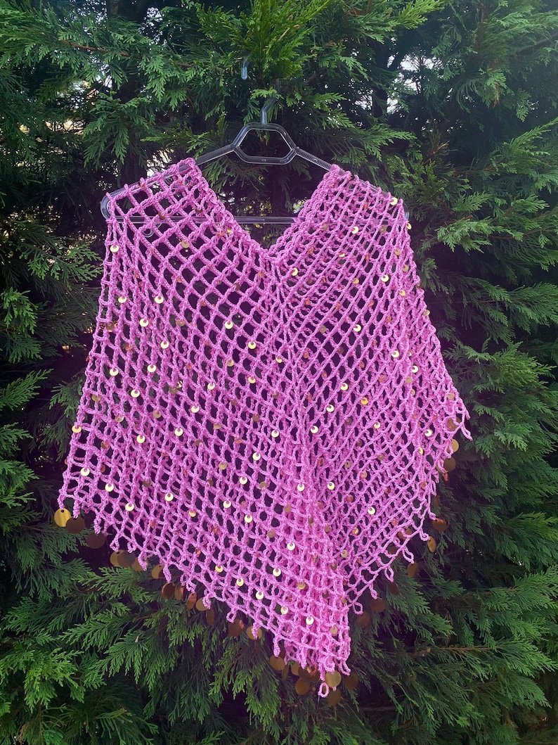 May include: A pink, open-weave crochet poncho with a V-neck. The poncho is adorned with gold-colored sequins throughout the body and along the bottom edge. The garment is displayed on a black hanger against a backdrop of green foliage.