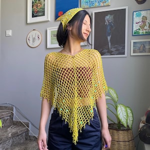 Puede incluir: Un poncho amarillo tejido a crochet con un diseño de red, adornado con pequeñas cuentas verde claro. La modelo lleva un pañuelo amarillo a juego, una camiseta granate y pantalones azul marino. El estilo general es bohemio.