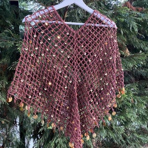 Può includere: Un poncho all'uncinetto bordeaux con scollo a V, ornato di paillettes dorate. La trama aperta è visibile e i bordi sono rifiniti con paillettes dorate pendenti. Il poncho è esposto su una gruccia bianca.