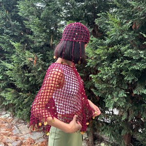 May include: A burgundy crochet hat and shawl set. The open-weave design is adorned with gold-colored sequins. The hat has dangling strands with sequins. The shawl is draped over a white top and green skirt, set against a backdrop of green foliage.