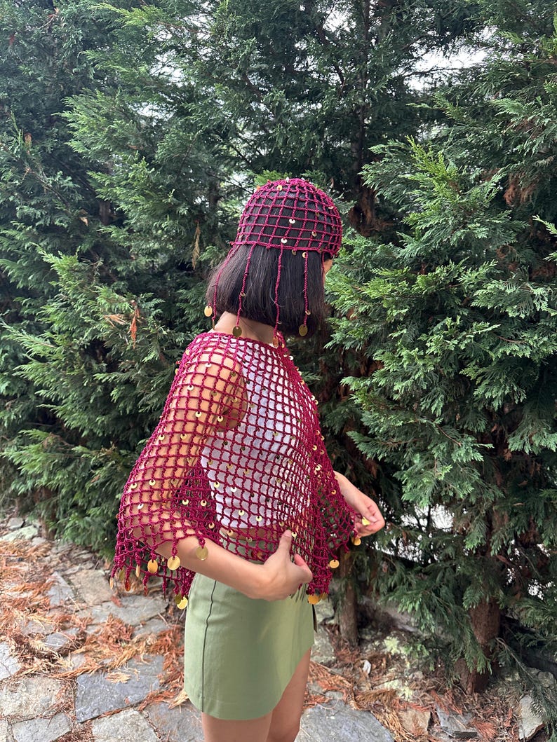 May include: A burgundy crochet hat and shawl set. The open-weave design is adorned with gold-colored sequins. The hat has dangling strands with sequins. The shawl is draped over a white top and green skirt, set against a backdrop of green foliage.