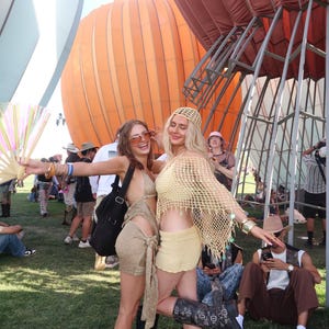 May include: Two people in festival attire pose outdoors. One wears a gold outfit with black boots, the other a yellow crochet ensemble with boots. Large orange and white hot air balloons are in the background.