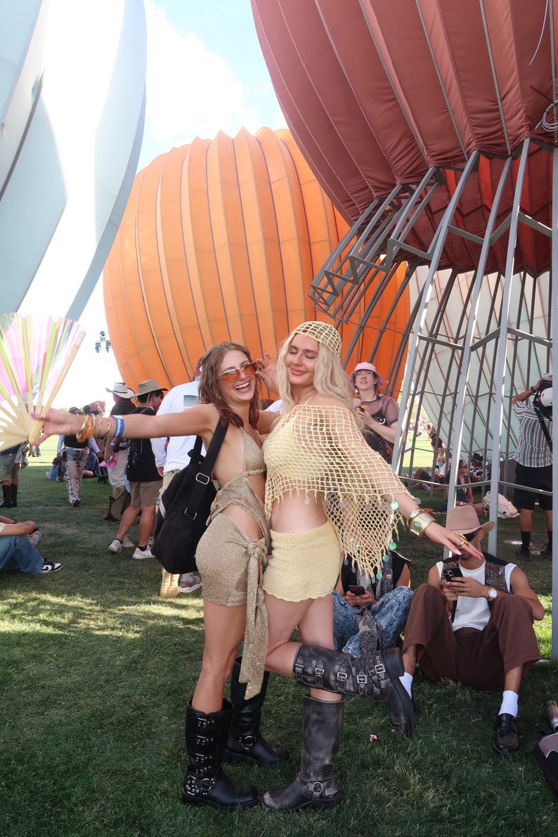 May include: Two people in festival attire pose outdoors. One wears a gold outfit with black boots, the other a yellow crochet ensemble with boots. Large orange and white hot air balloons are in the background.