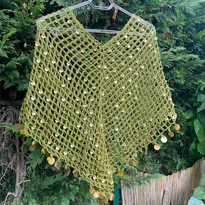 Könnte beinhalten: Ein grüner gehäkelter Poncho mit V-Ausschnitt, verziert mit goldfarbenen Pailletten. Das offene Webmuster und der Paillettenfransen erzeugen einen dekorativen, böhmischen Stil. Der Poncho wird auf einem Kleiderbügel vor grünem Laub gezeigt.