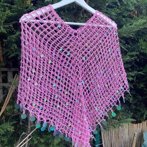 May include: A pink, open-weave crochet poncho with iridescent sequins. The poncho is displayed on a white hanger against a backdrop of green foliage. The sequins are round and add a touch of sparkle to the garment.
