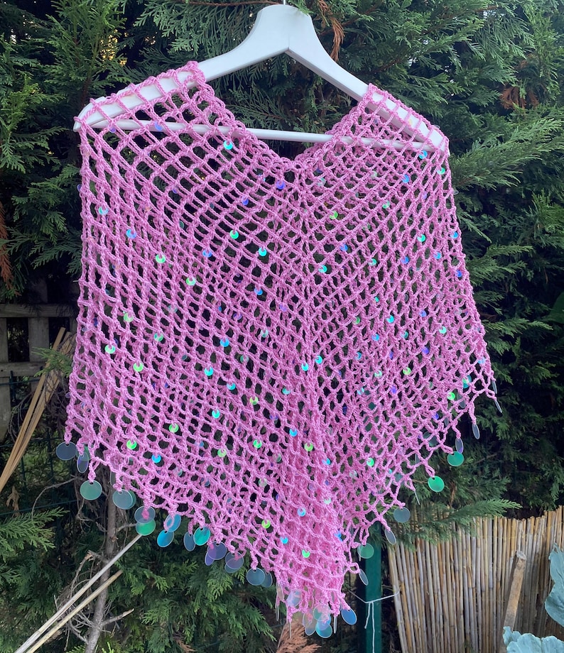 May include: A pink, open-weave crochet poncho with iridescent sequins. The poncho is displayed on a white hanger against a backdrop of green foliage. The sequins are round and add a touch of sparkle to the garment.