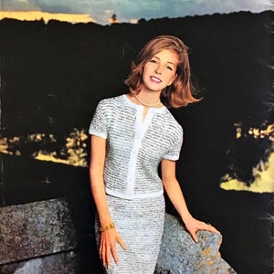 May include: A woman wearing a two-piece knit outfit. The outfit includes a short-sleeved, button-down top and a knee-length skirt. The knitwear is white with a subtle black pattern. The background features a sunset over a dark landscape.
