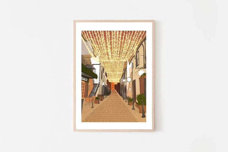 May include: A narrow, cobblestone street lined with buildings, decorated with twinkling string lights overhead. The street is empty, with a few potted plants and a sign that reads "Cafe" visible.