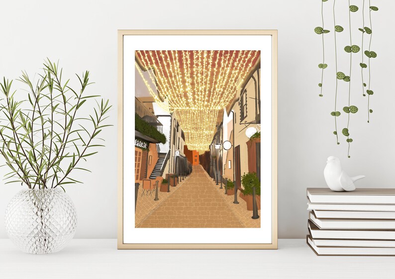 May include: A digital illustration of a narrow street lined with buildings, decorated with string lights. The street is paved with brick and the buildings are painted in warm tones.