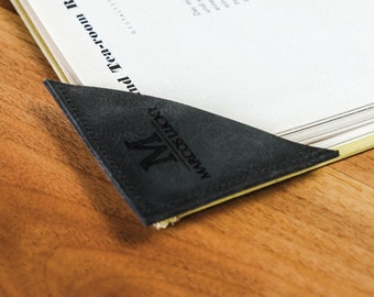 May include: A black leather journal with a pen holder and a bookmark. The journal has a gold metal clasp and the bookmark has the text "MARCOS LUKKY" and a letter "M" on it. The journal is open to a page with the text "and Tea-room Ritz in Basel".