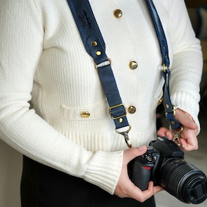 May include: A person wearing a white sweater with gold buttons and a blue leather camera strap with the word "Taltiu" embossed on it. The person is holding a black DSLR camera with a large lens.