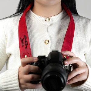 May include: A red leather camera strap with the name "Julia" engraved on it. The strap is being worn by a person holding a black DSLR camera.