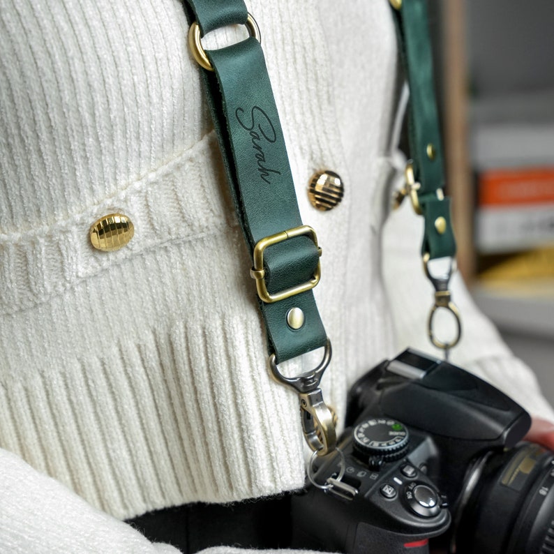 May include: A green leather camera strap with gold hardware and the name "Sarah" engraved on it. The strap is attached to a black DSLR camera.