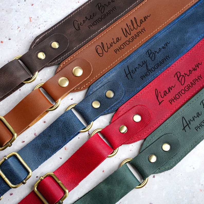 May include: Five leather camera straps in different colours, each with a personalised name. The straps are brown, blue, red, green and black. Each strap has two brass metal rings and two brass metal buttons. The names on the straps are George Brown Photography, Olivia William Photography, Henry Brown Photography, Liam Brown Photography and Anna Thomas Photography.