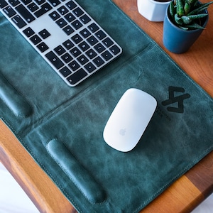 May include: A teal leather desk mat with a white wireless mouse and a silver keyboard. The mat has a stitched edge and a leather wrist rest. The mat has the text "Developer Flex" and a logo printed on it.