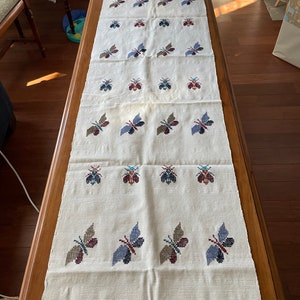 K&ouml;nnte beinhalten: Ein wei&szlig;er Tischl&auml;ufer mit einem sich wiederholenden Muster aus gestickten Schmetterlingen in Blaut&ouml;nen, Rot und Braun. Die Schmetterlinge sind in Reihen angeordnet und gleichm&auml;&szlig;ig verteilt.
