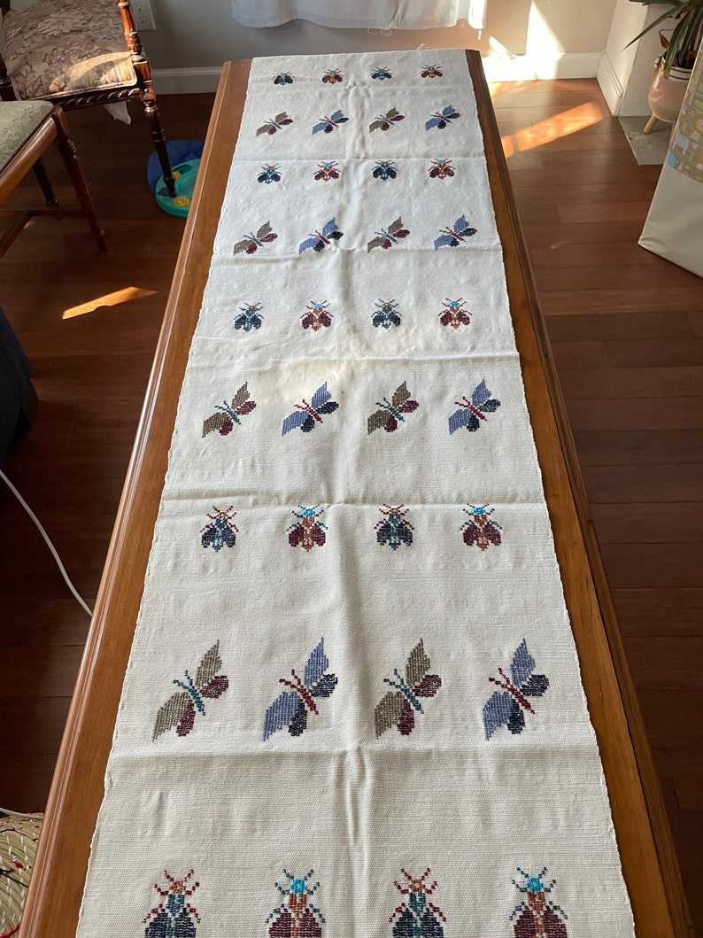 K&ouml;nnte beinhalten: Ein wei&szlig;er Tischl&auml;ufer mit einem sich wiederholenden Muster aus gestickten Schmetterlingen in Blaut&ouml;nen, Rot und Braun. Die Schmetterlinge sind in Reihen angeordnet und gleichm&auml;&szlig;ig verteilt.